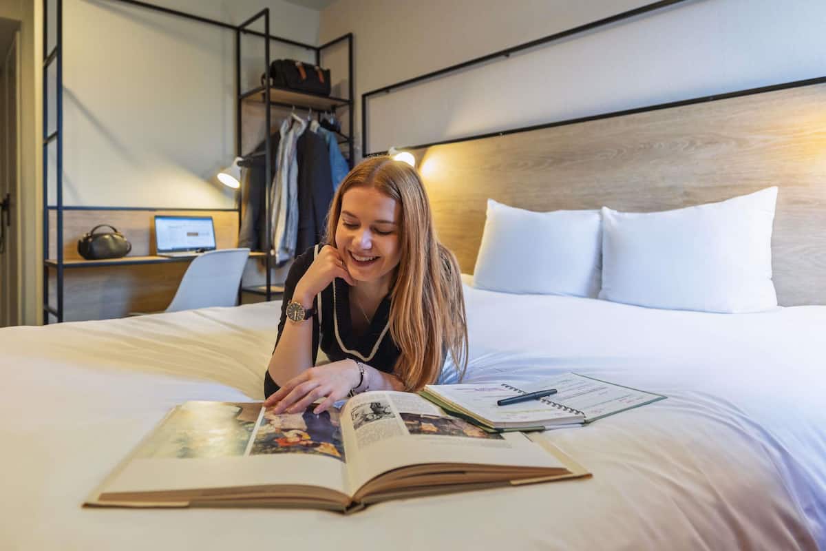 Quarto de hotel com uma cama, uma escrivaninha com um laptop, uma estante e uma prateleira fixada na parede com roupas.