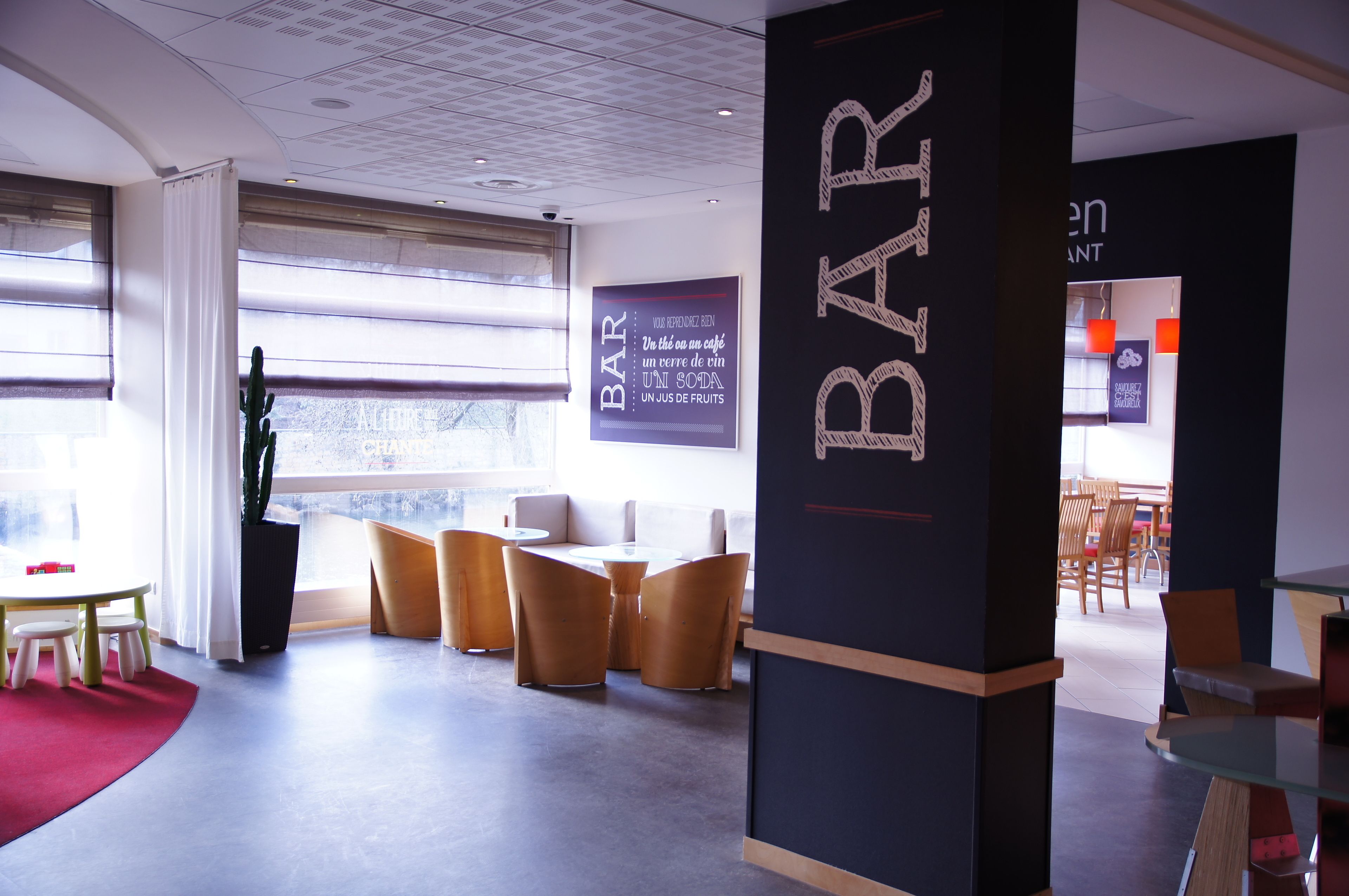 Een moderne barruimte met een groot bord waarop 'BAR' staat en een slogan in een andere taal. Er zijn verschillende houten stoelen en tafels, en een potplant is zichtbaar.