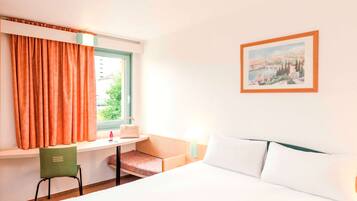 Une chambre d’hôtel avec un lit, un bureau, une chaise, une fenêtre avec des rideaux et un tableau au mur.