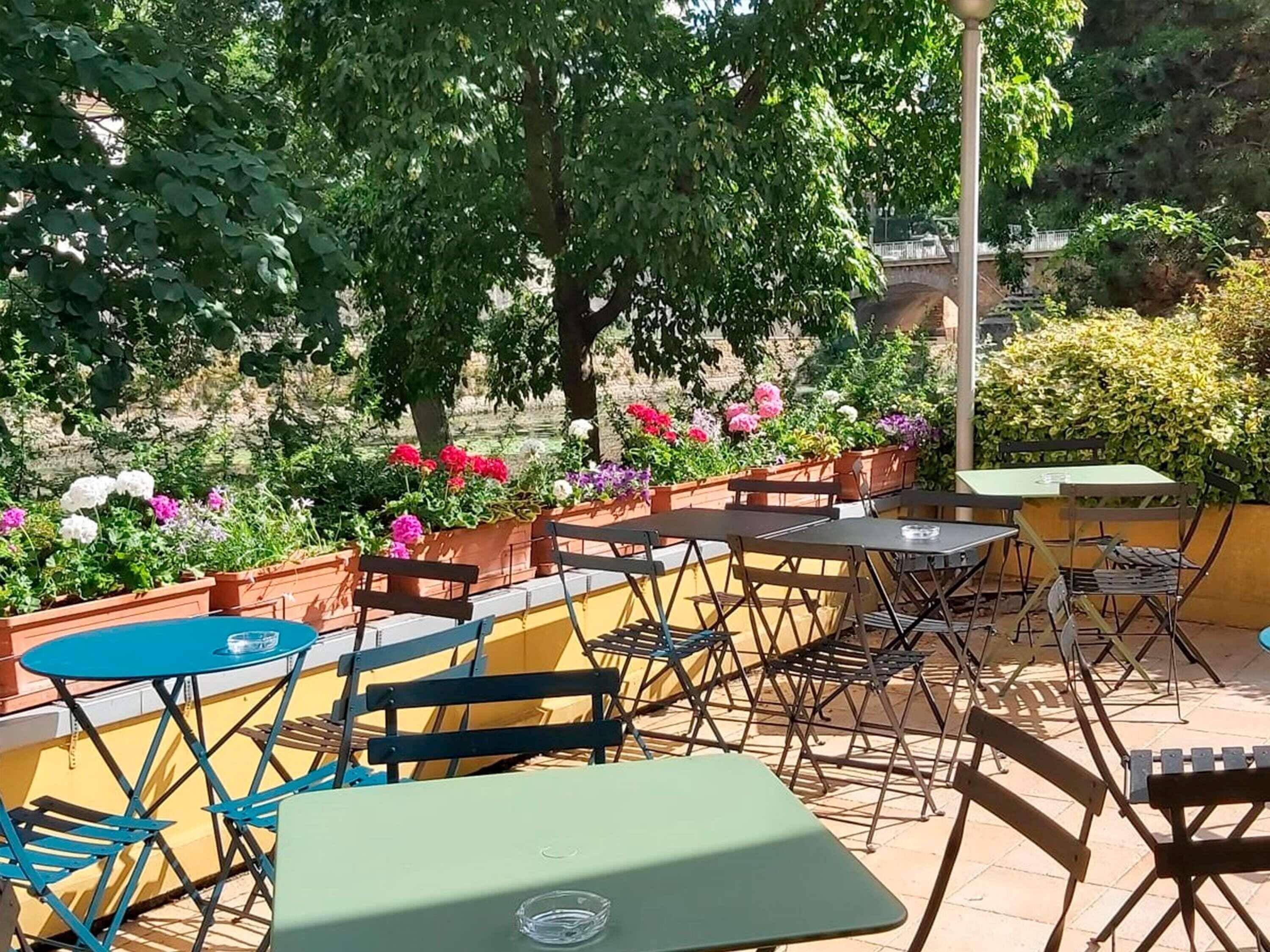An outdoor seating area with tables and chairs, surrounded by flower beds and trees.