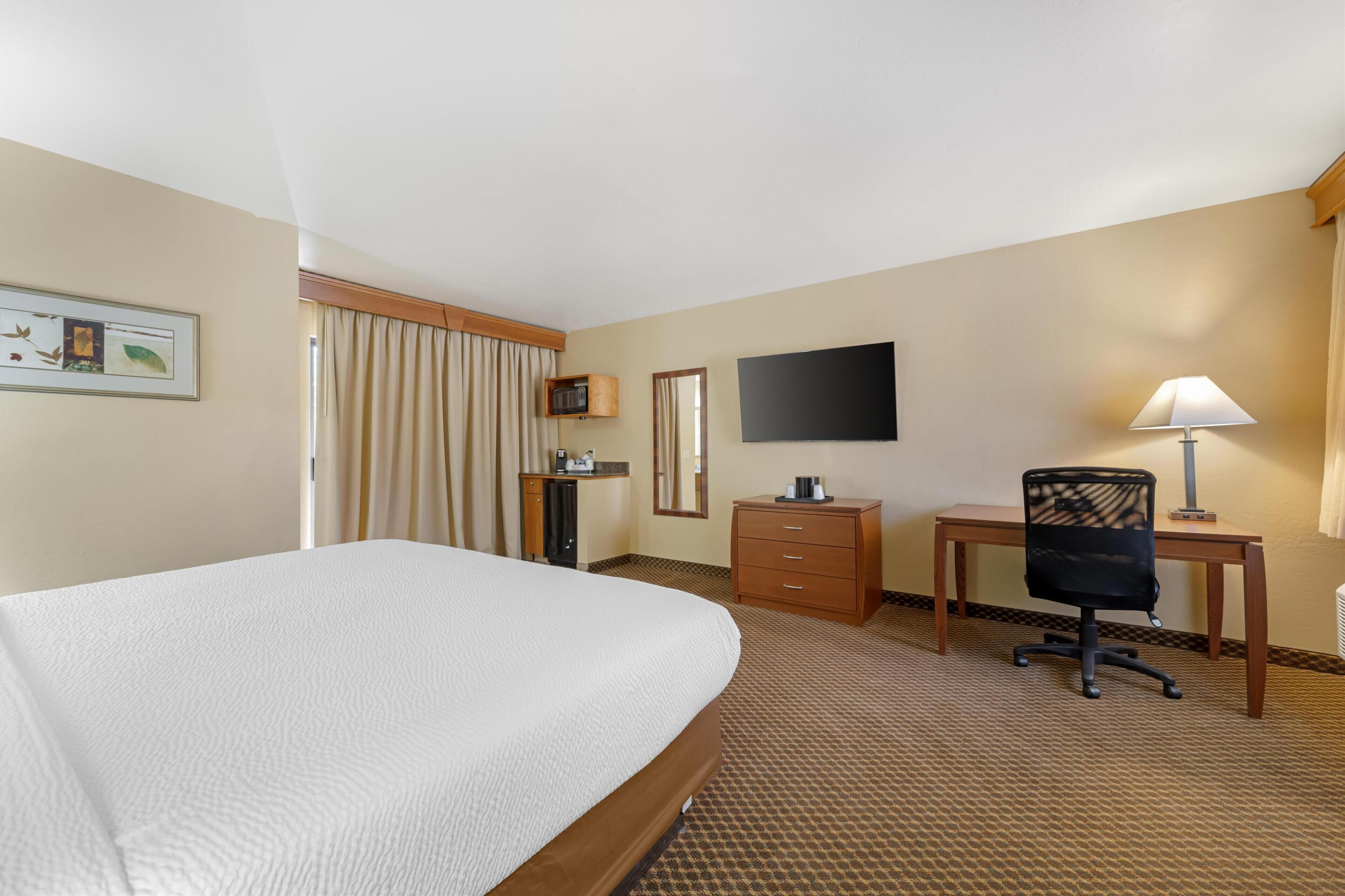 A hotel room with a large bed, a desk with a chair, a television, a lamp, and a small cabinet.