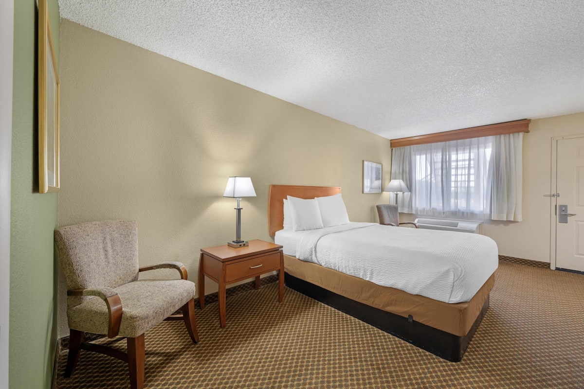 A hotel room with a bed, a chair, a nightstand, and a window with curtains.