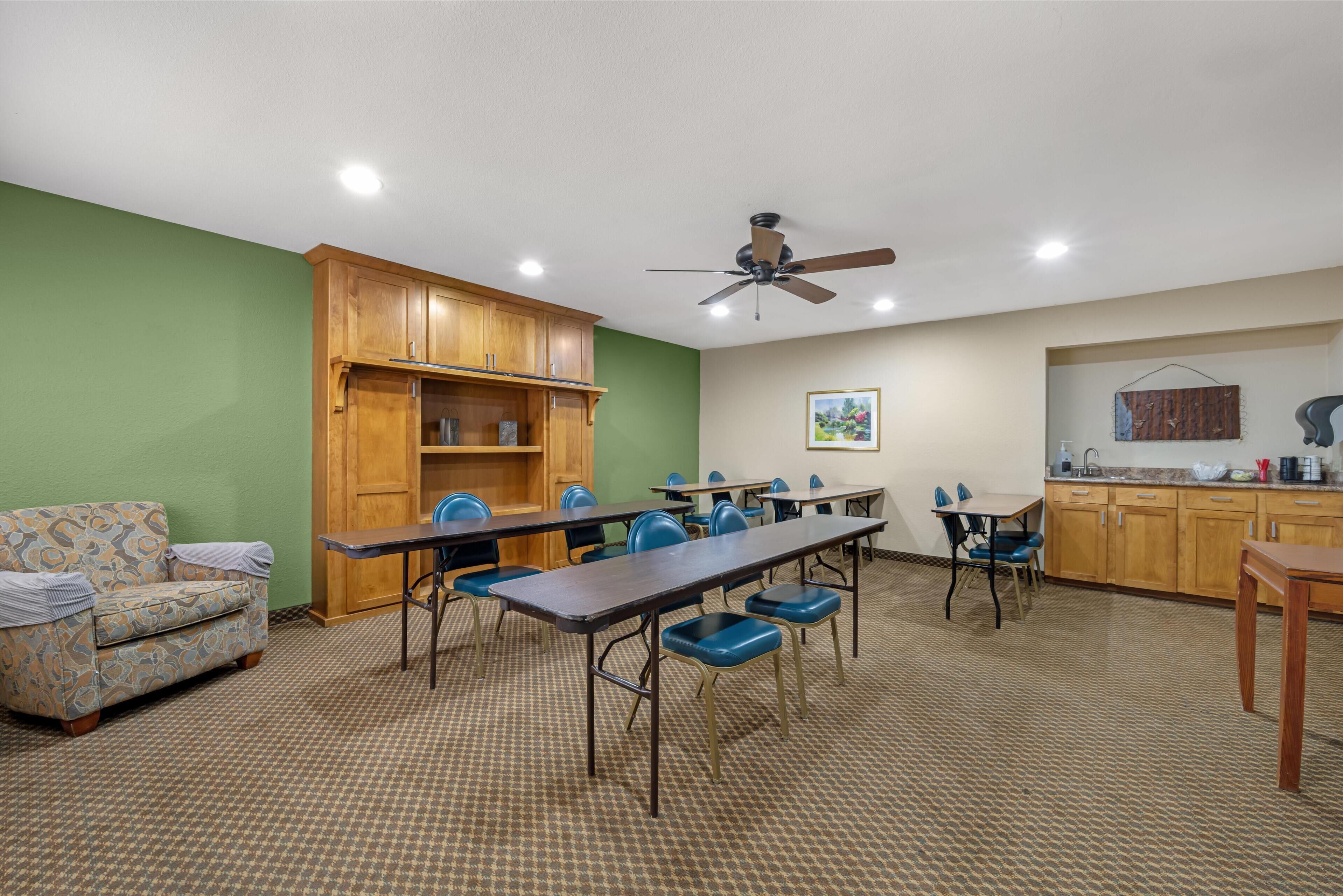 A room with a long table, blue chairs, a wooden cabinet, a ceiling fan, and a kitchenette.
