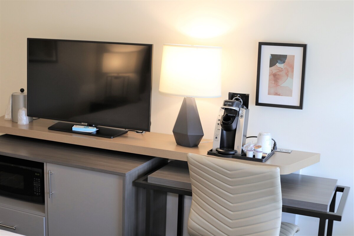 A modern hotel room with a flat-screen TV, a coffee machine, a lamp, and a framed picture on the wall.