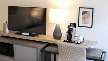 A hotel room with a flat-screen TV, a coffee machine, a lamp, and a framed picture on the wall.