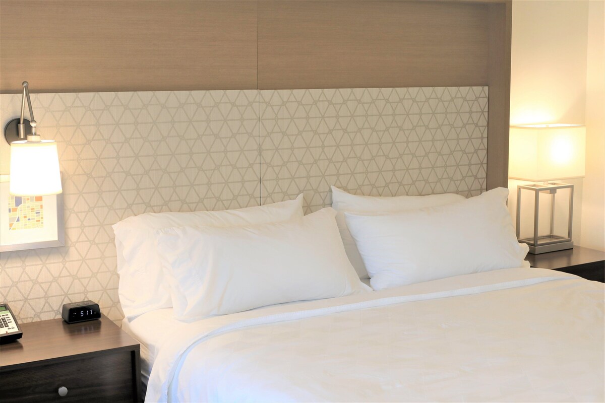 A neatly made bed with white linens, a bedside table with a lamp, and a wall clock.