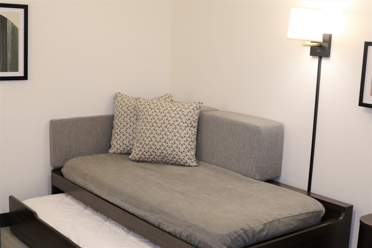 A single bed with a grey mattress and patterned pillows, a bedside table, and a wall-mounted lamp.