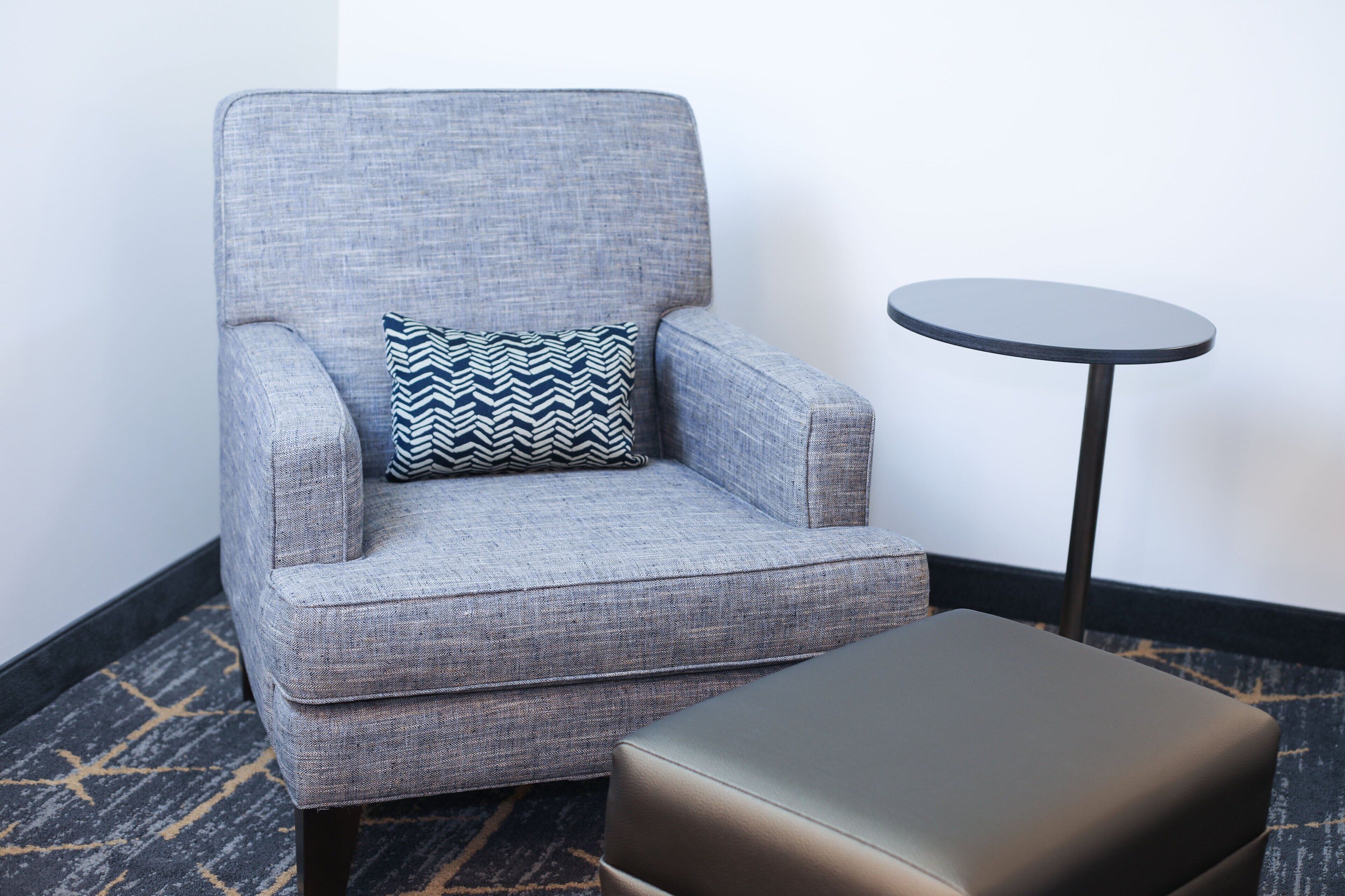 A grey armchair with a patterned pillow, a small round table, and a matching ottoman.