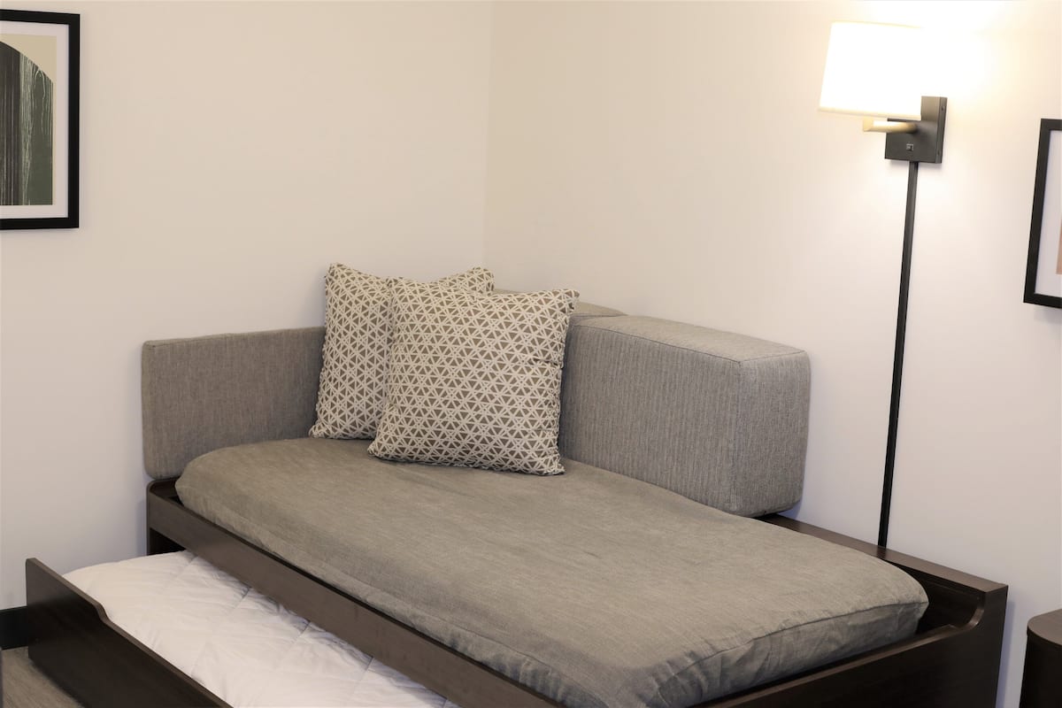 A single bed with a grey mattress and patterned pillows, a bedside table, and a wall-mounted lamp.