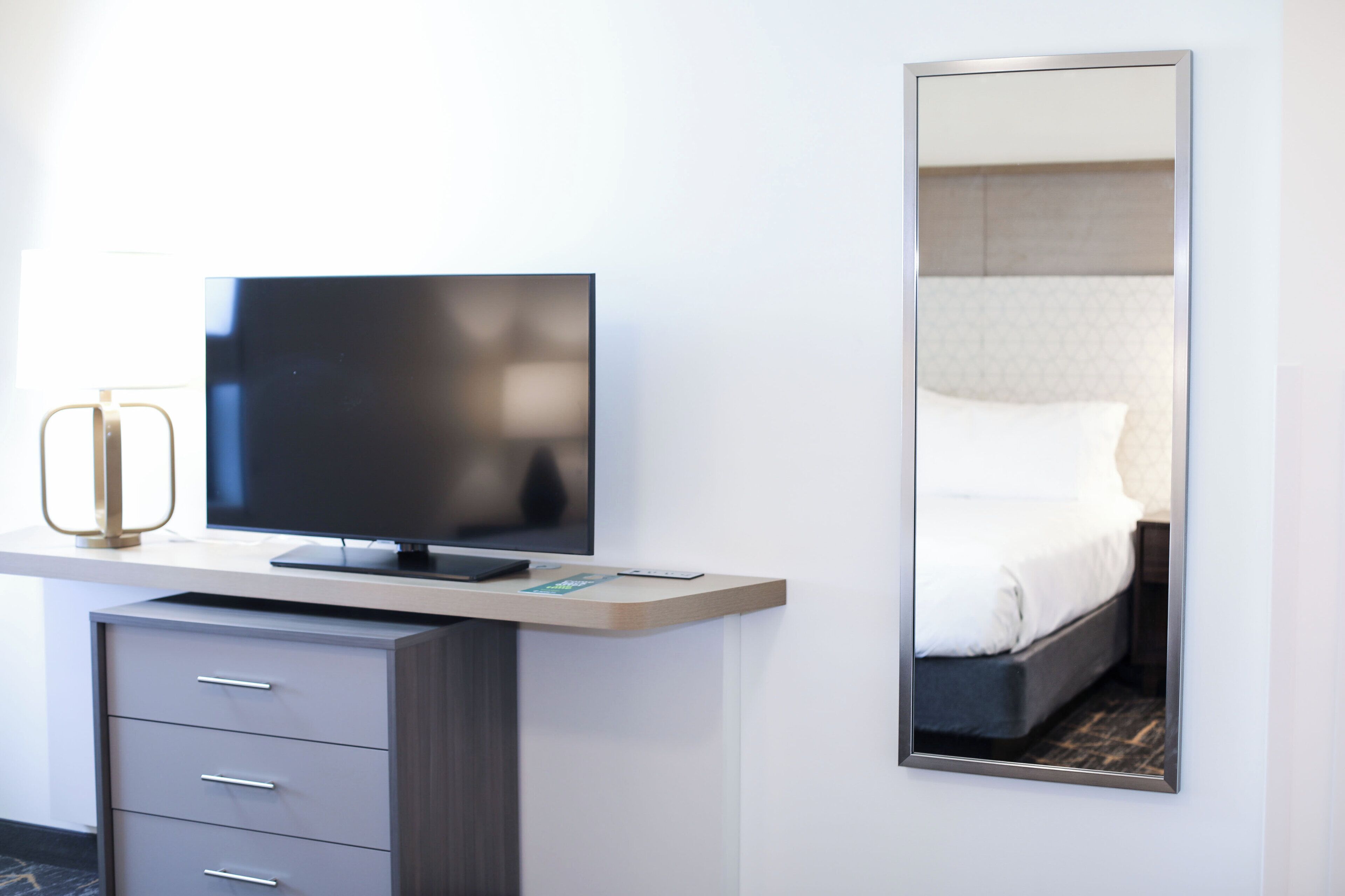 A hotel room with a flat-screen TV on a wooden console, a mirror reflecting the bed, and a built-in desk with drawers.