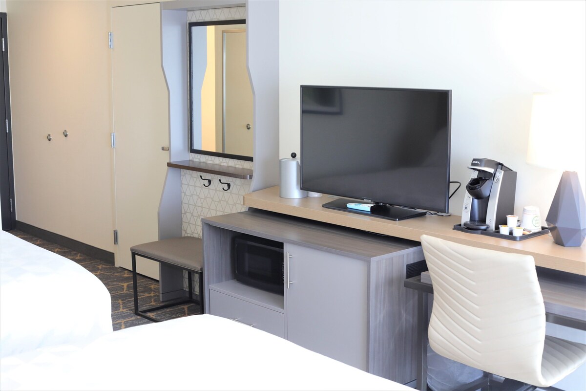 A hotel room with a bed, a desk with a computer, a chair, a microwave, and a coffee maker.