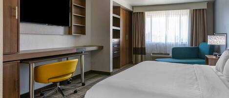 A hotel room with a bed, a desk with a chair, a television, a wardrobe, and a window with curtains.