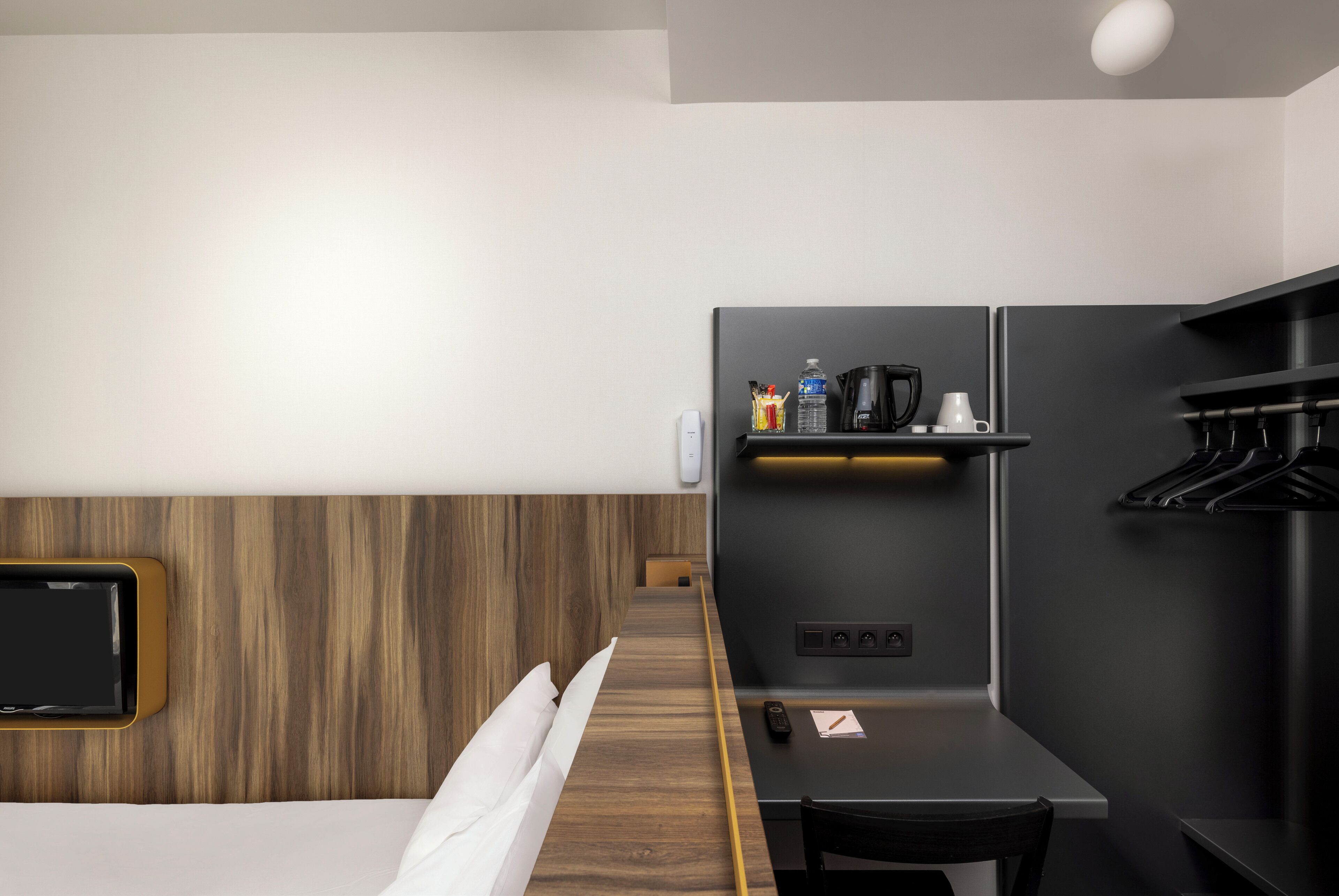 Una habitación de hotel moderna con cabecera de madera, una cama, un escritorio con silla, un televisor de pantalla plana, una cafetera y un estante pequeño con artículos de aseo.