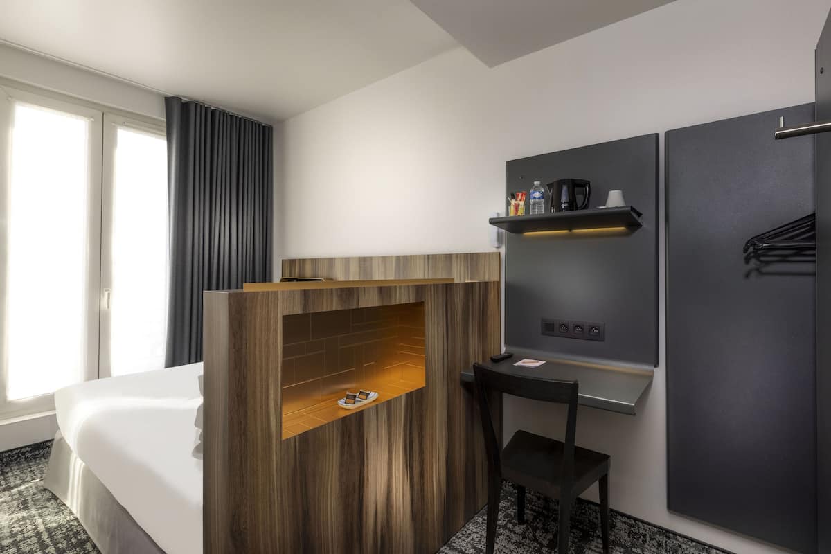 Hotelkamer met een bed, een houten plank met een schoen, een klein tafeltje met een stoel, een wandplank met toiletartikelen en een donkerhouten kast.