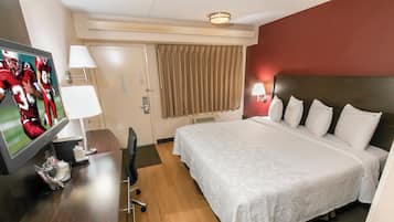 Habitación de hotel con cama, escritorio con televisor, silla, lámpara y ventana con cortinas.