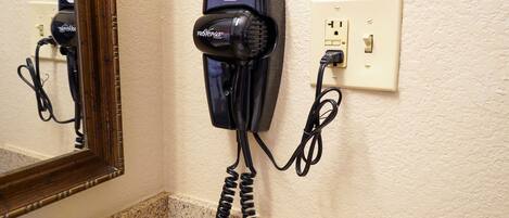 A hair dryer mounted on a wall with a power outlet and a coiled cord.