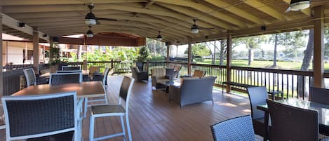 An outdoor covered patio with wooden flooring, white and dark wicker furniture, ceiling fans, and potted plants.