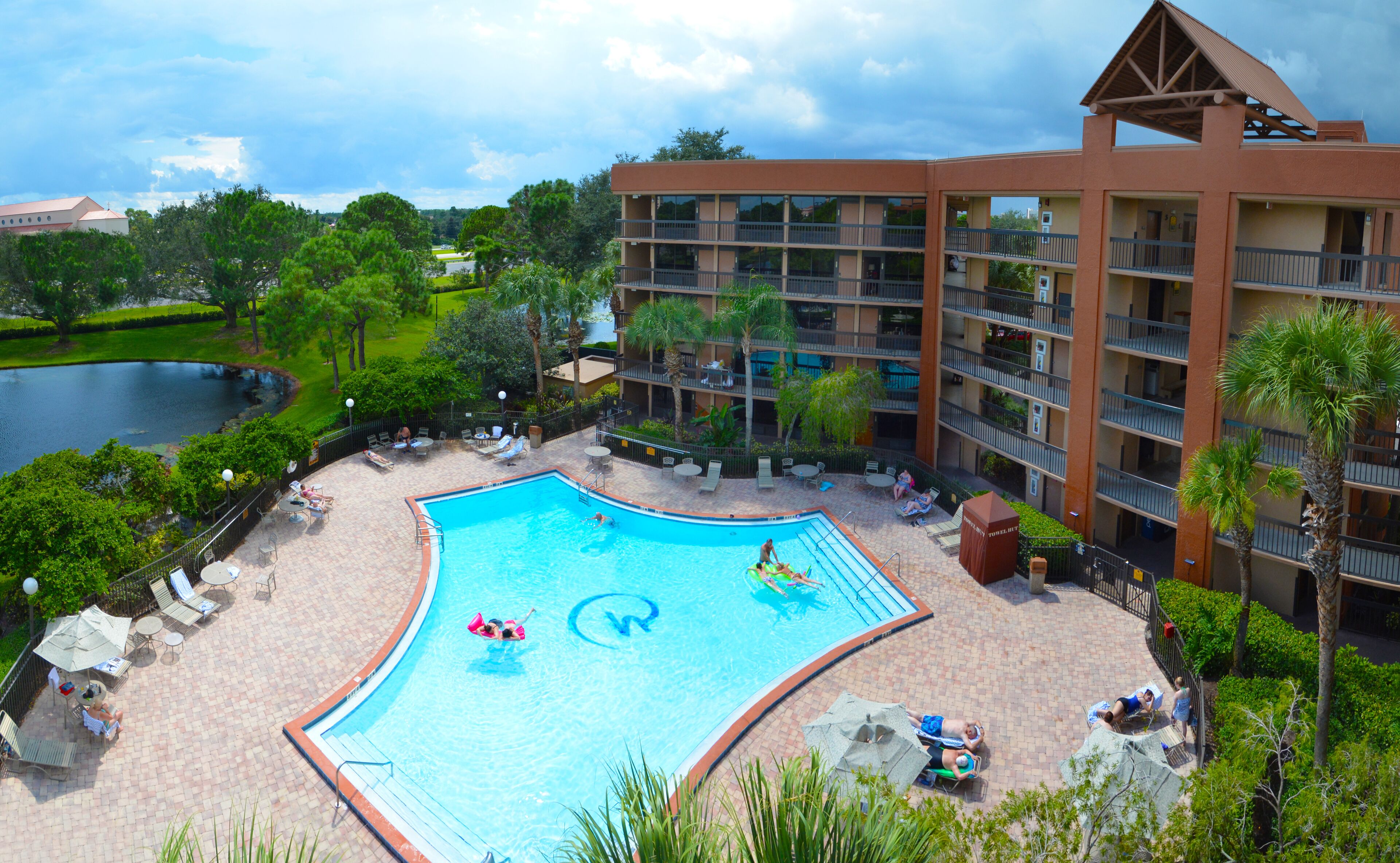 Um hotel com piscina, cercada por espreguiçadeiras e guarda-sóis, quadra de tênis e vista para um lago.