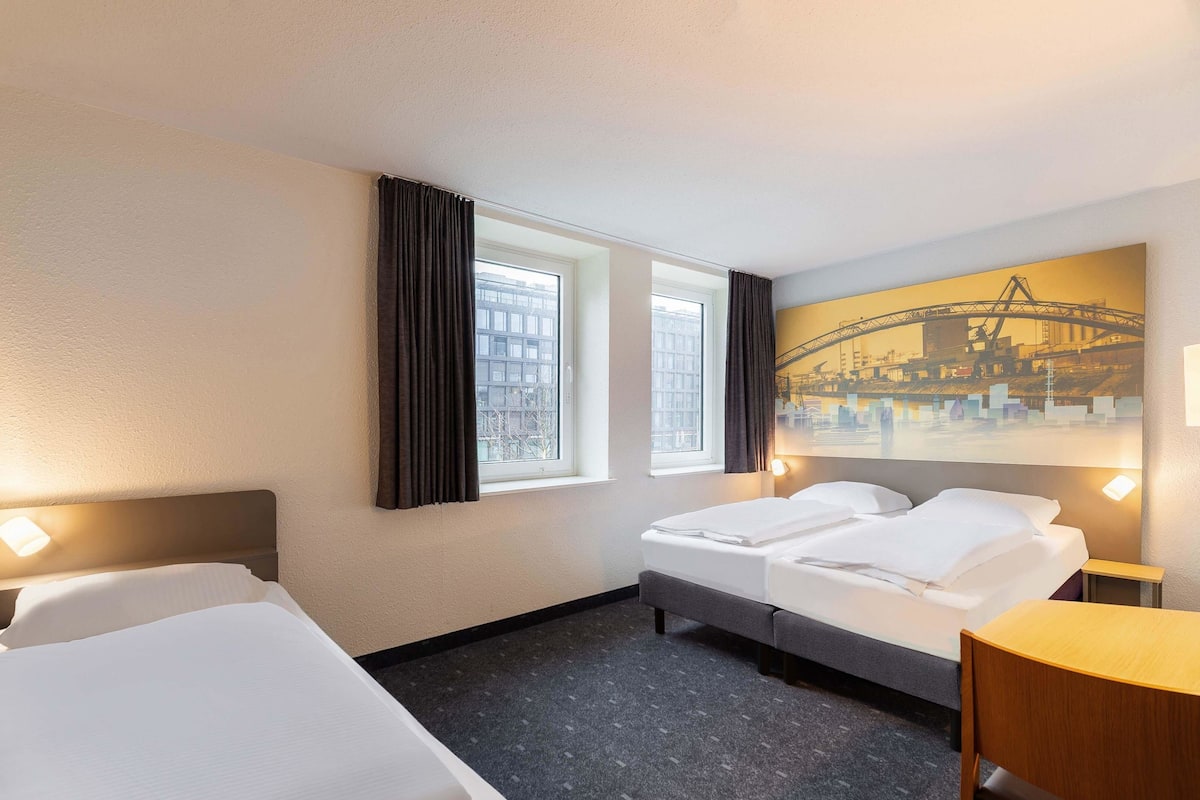 Une chambre d’hôtel avec deux lits, un bureau et une grande fenêtre avec des rideaux.