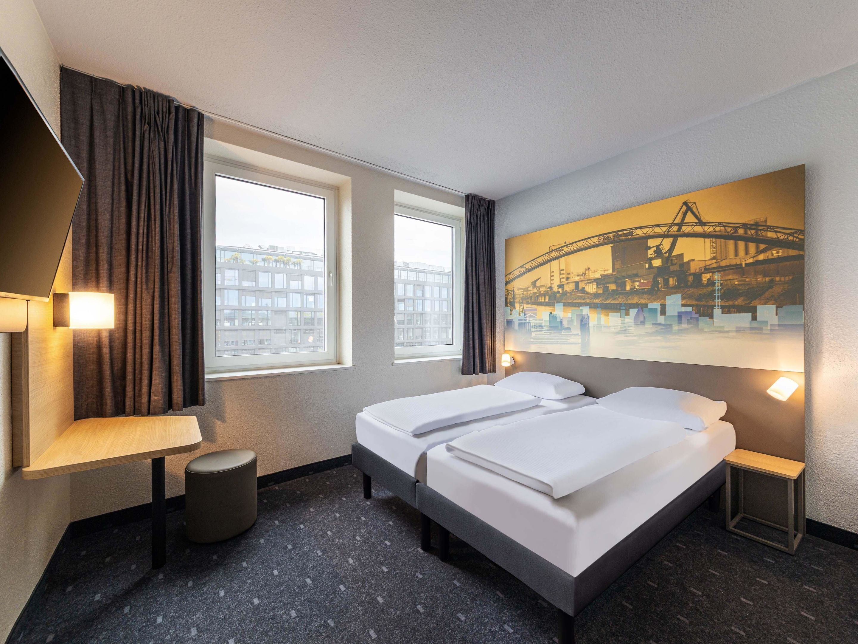 A hotel room with two beds, a desk, a chair, a small stool, and a large window with curtains.