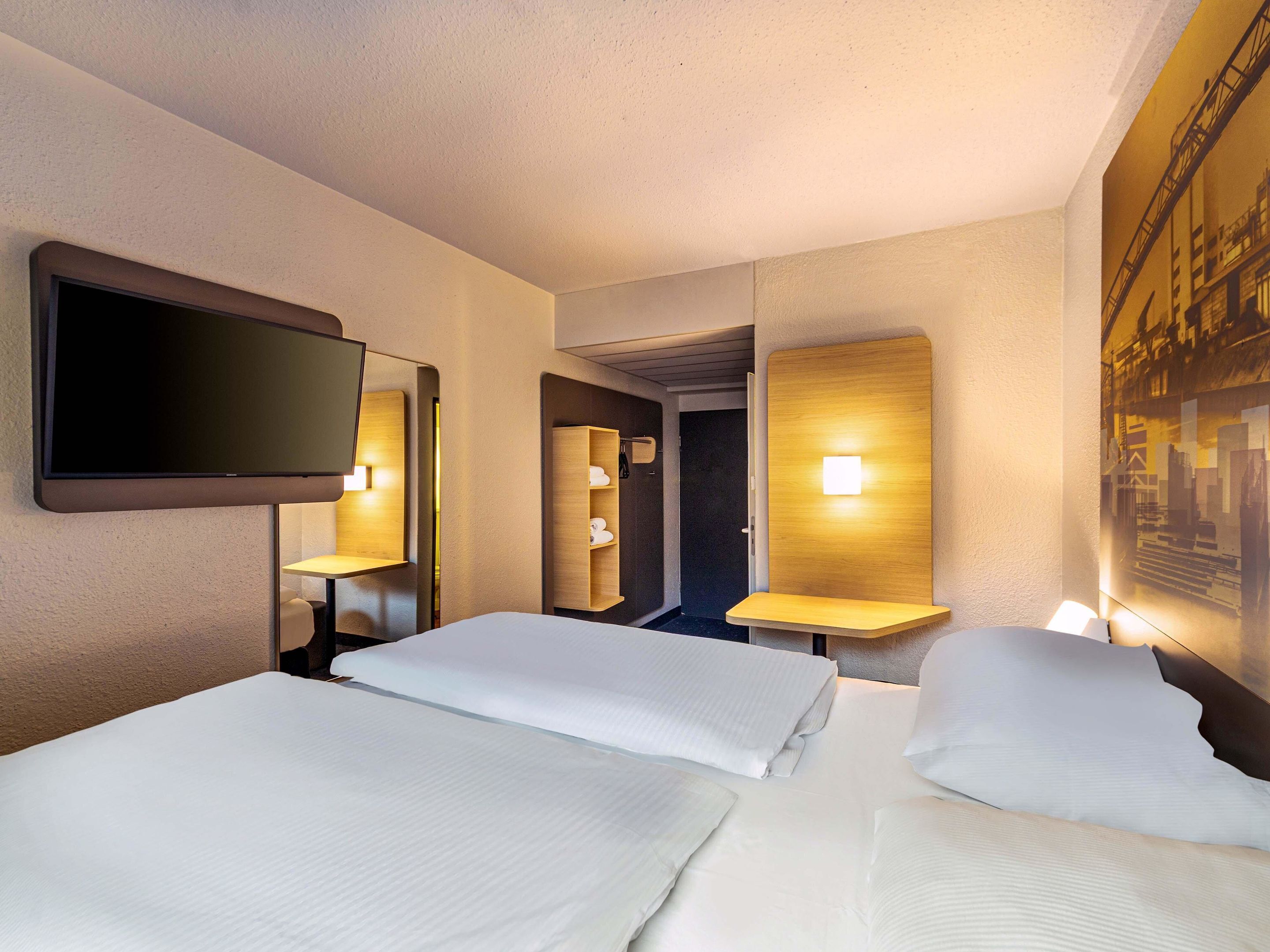 Quarto de hotel com duas camas, uma televisão de tela plana na parede e um quadro de uma cena industrial.