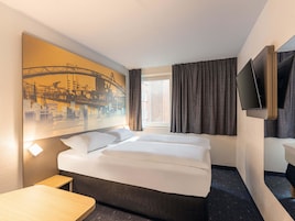 Quarto de hotel com uma cama grande, uma escrivaninha, uma TV de tela plana na parede e uma janela com cortinas.