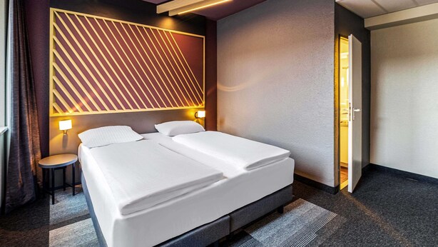 Een moderne hotelkamer met een groot bed, twee nachtkastjes, een wandlamp en een deur naar een andere kamer.