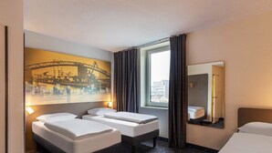 Een hotelkamer met twee bedden, een groot raam met gordijnen, een stadslandschap schilderij aan de muur en een spiegel.