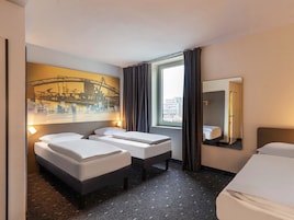 Quarto de hotel com duas camas, uma janela grande com cortinas, um quadro de paisagem urbana na parede e um espelho.
