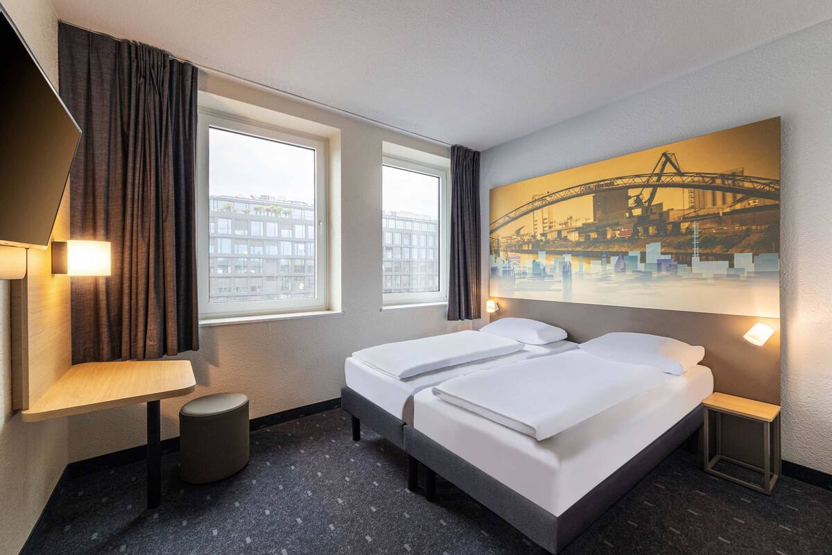 Een hotelkamer met twee bedden, een bureau, een stoel, een klein krukje en een groot raam met gordijnen.