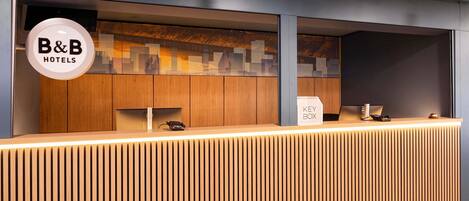 A modern reception desk with a wooden finish and a glass partition. A key box and a phone are visible on the desk.