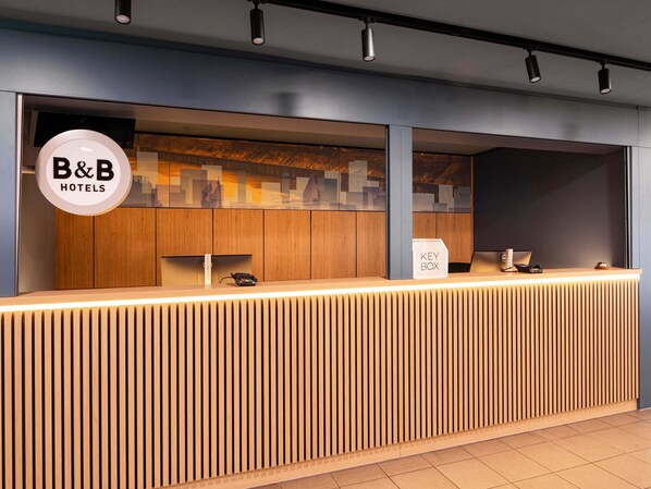 A modern reception desk with a wooden finish and a glass partition. A key box and a phone are visible on the desk.