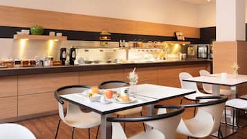Uma sala de jantar moderna com mesas e cadeiras brancas, um balcão de buffet com diversas opções de comida e uma geladeira ao fundo.