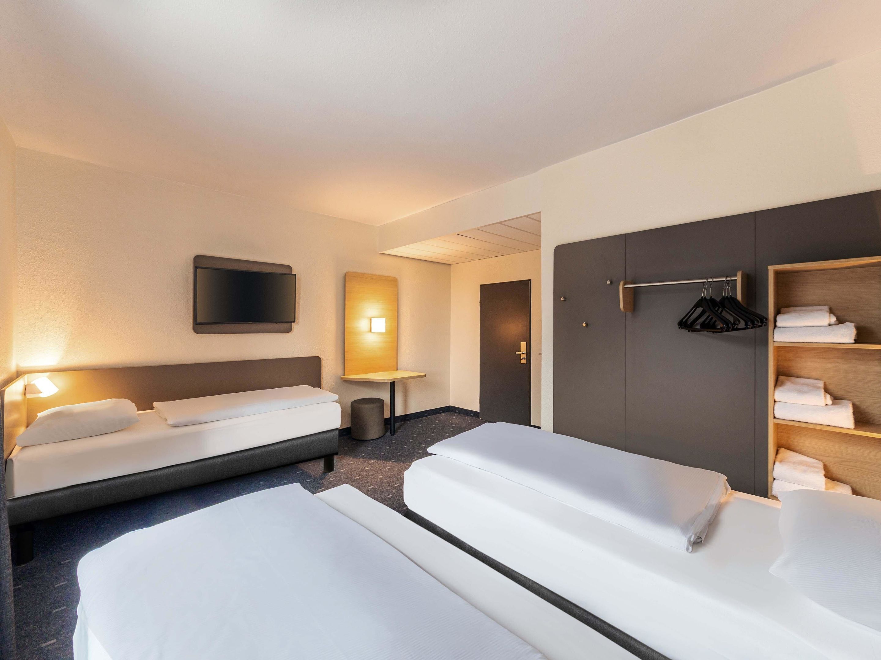 Habitación de hotel con dos camas, televisión, mesita de noche, espejo y armario con ropa colgada.