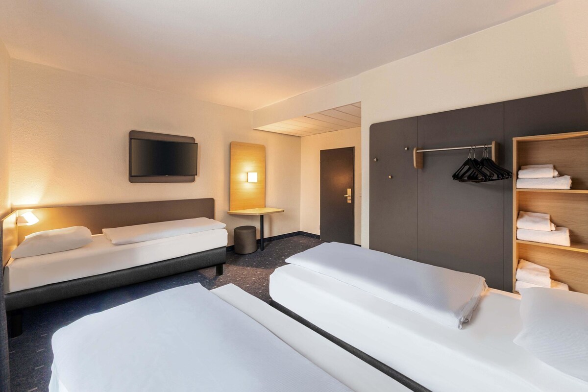 Une chambre d’hôtel avec deux lits, une télévision, une table de chevet, un miroir et une armoire avec des vêtements suspendus.
