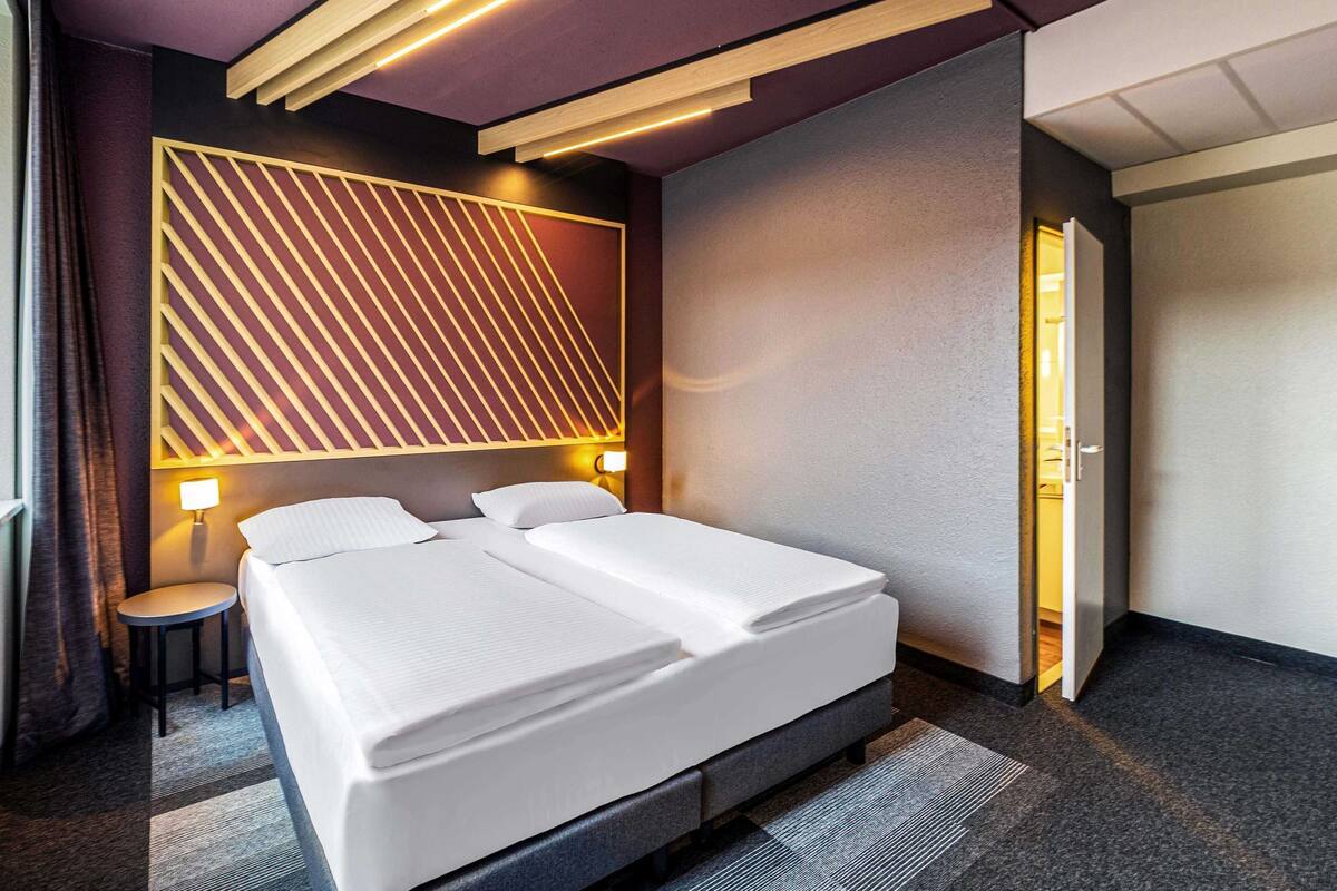 Een moderne hotelkamer met een groot bed, twee nachtkastjes, een wandlamp en een deur naar een andere kamer.