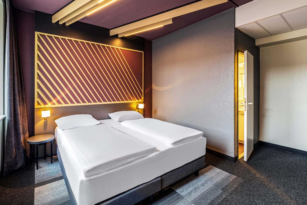 Une chambre d’hôtel moderne dotée d’un grand lit, de deux tables de chevet, d’un panneau lumineux fixé au mur et d’une porte donnant sur une autre pièce.