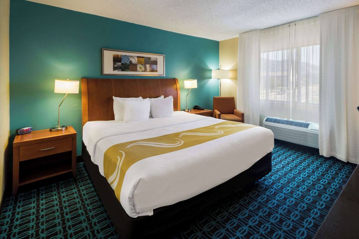 A hotel room with a large bed, two bedside tables, a chair, a window with curtains, and a wall-mounted picture.