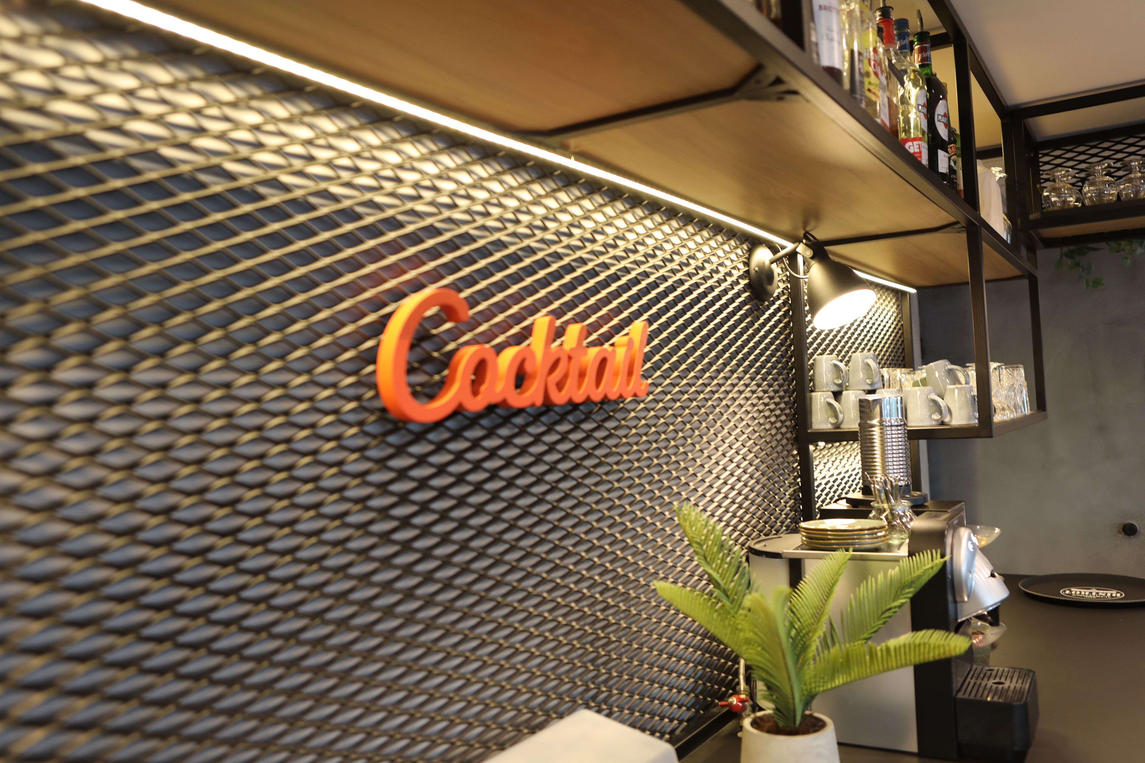 Área de bar com um letreiro 'Cocktail' em uma parede texturizada, uma planta e utensílios de bar nas prateleiras.