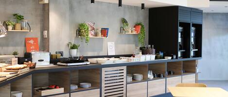 Un área de cocina moderna con una encimera larga, varios electrodomésticos y estantes con plantas y libros.
