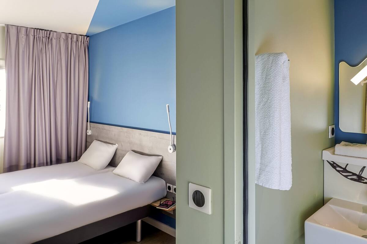 Habitación de hotel moderna con cama, mesita de noche, espejo y baño con lavabo y toalla.