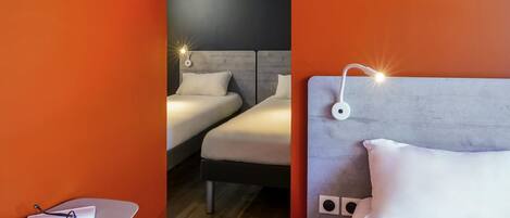 A hotel room with two beds, a small table with glasses and a book, and a wall-mounted speaker.