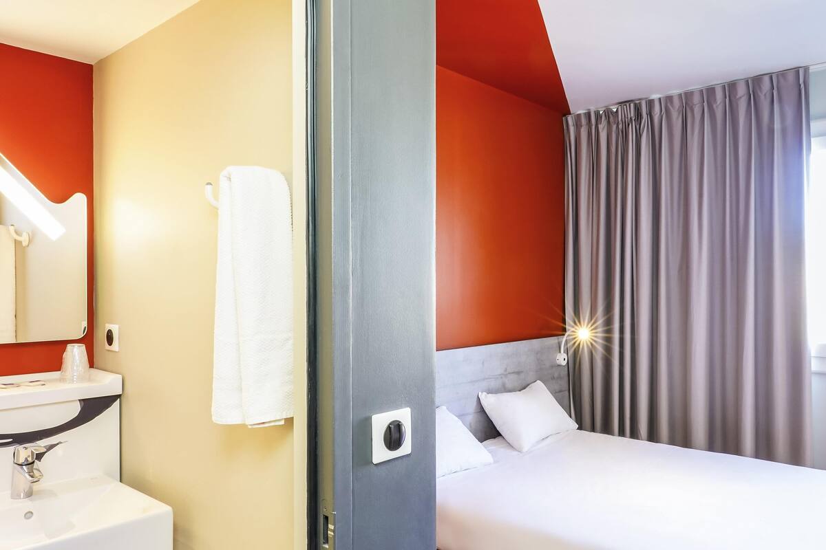 Una habitación de hotel con una cama, un lavamanos blanco, un espejo y una ventana con cortinas.