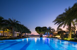 Piscina con sillones de descanso y palmeras, rodeada de edificios con ventanas iluminadas.