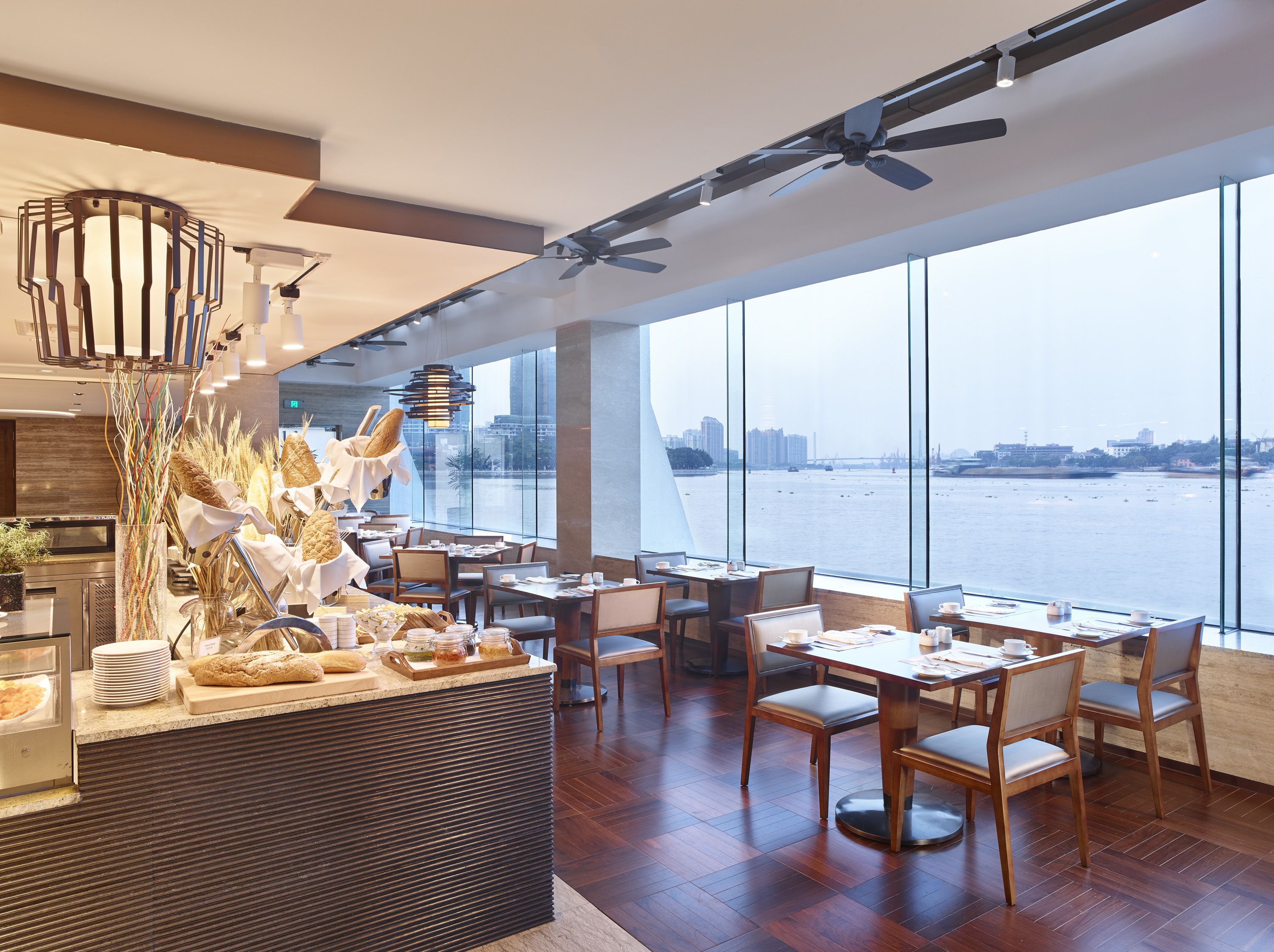 A modern restaurant with a view of the water, wooden flooring, and a buffet area with bread and pastries.