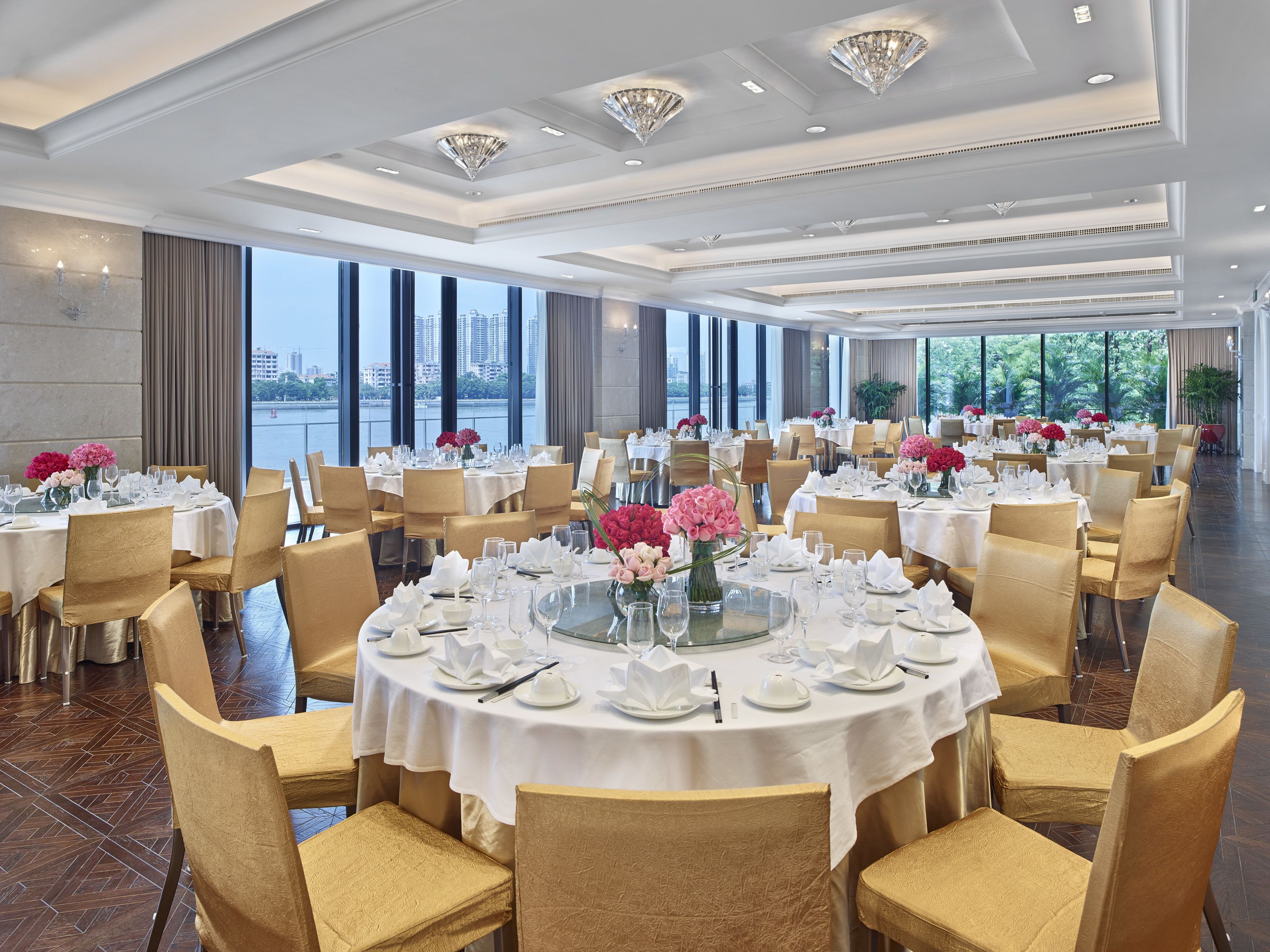 A spacious dining area with round tables set for a formal meal, large windows offering a view of a cityscape, and elegant chandeliers.