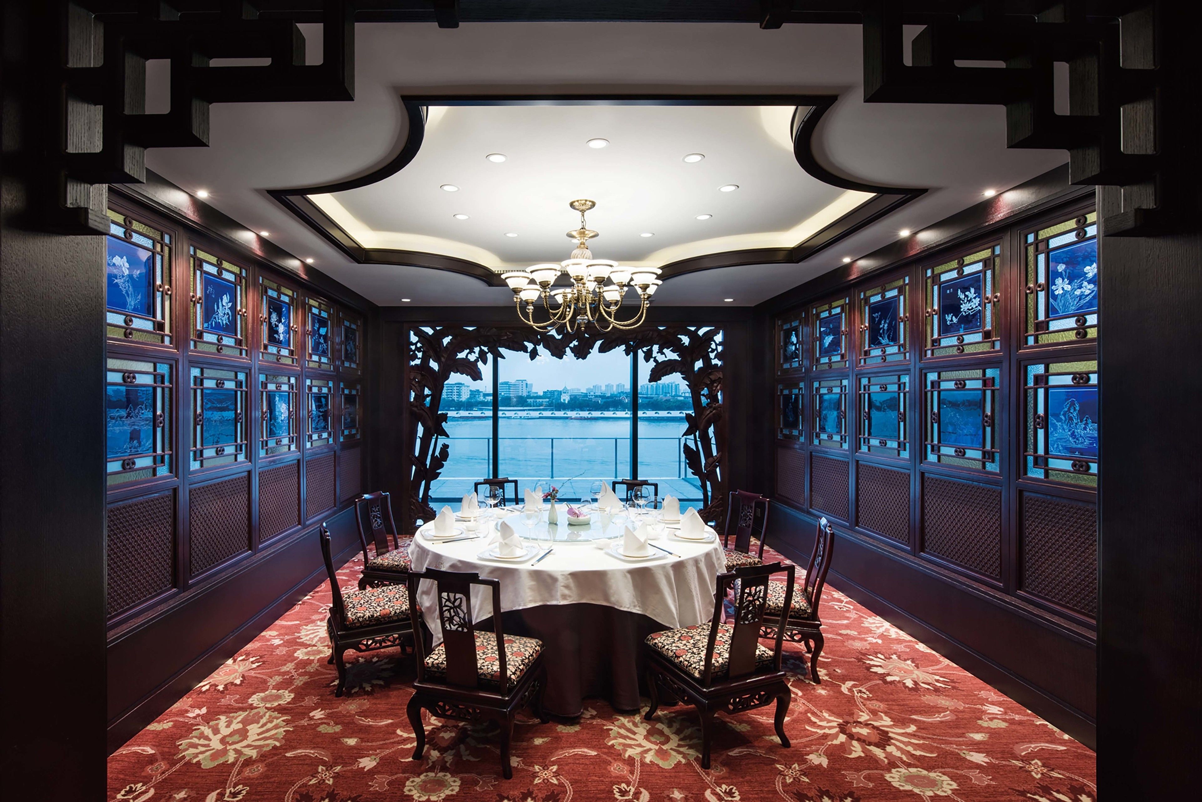 A dining area with a round table set for six, surrounded by chairs with floral upholstery. The room features a large chandelier, ornate wooden paneling, and large windows with decorative glass panes.