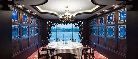 A dining area with a round table set for six, surrounded by chairs with floral upholstery. The room features a large chandelier, ornate wooden paneling, and large windows with decorative glass panes.