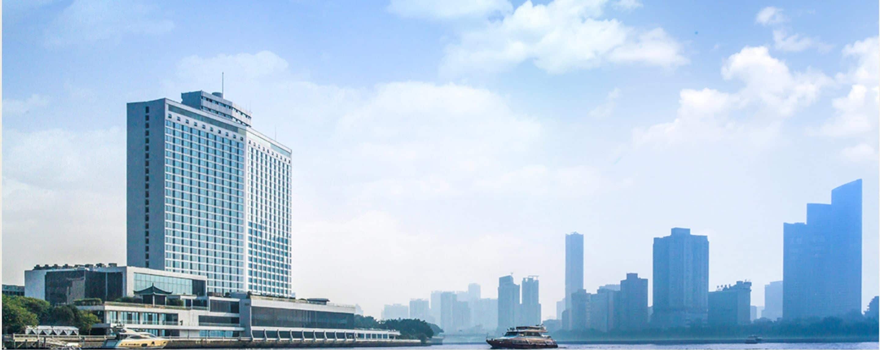A high-rise hotel with a modern glass facade, situated by a waterfront with a view of the city skyline.