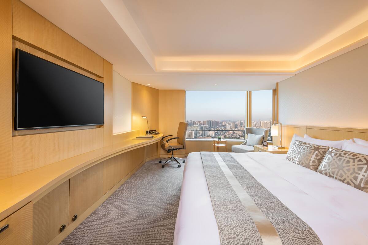 A hotel room with a large bed, a desk with a chair, a television, and a city view.